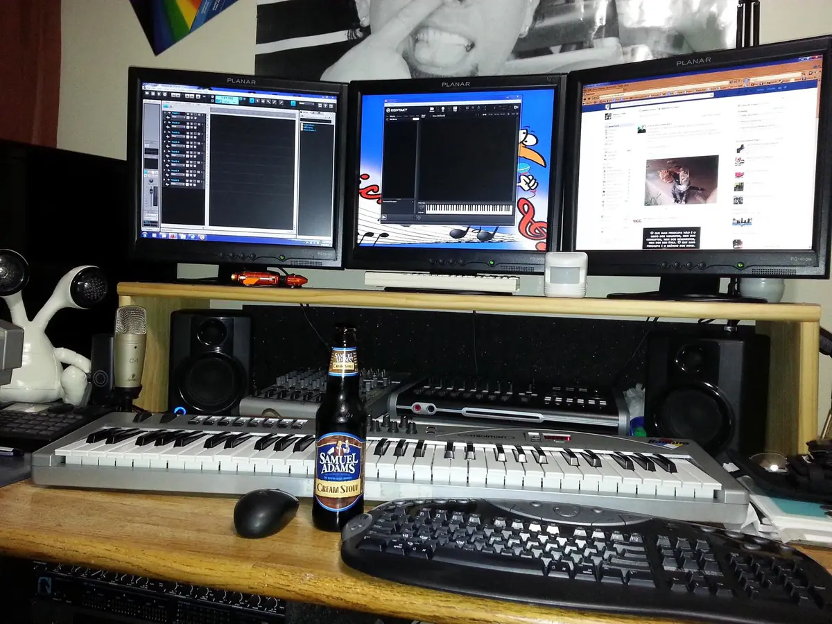 Three-monitor desk with the DAW on screen, keyboard, mouse, headphones and a cold one