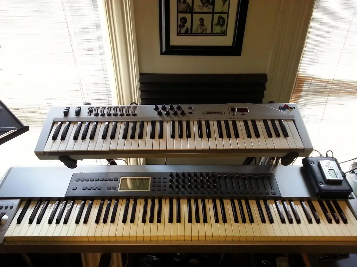 Two stacked MIDI keyboards and synthesizers on a table by a window with blinds