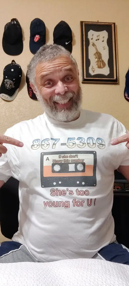 Angelo in a Journey 867-5309 cassette t-shirt pointing at the logo, caps hanging on the wall behind